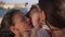 Close-up portrait of carefree pretty baby girl with cheerful sisters kissing toddler on cheeks in slow motion. Happy