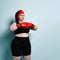 Close up portrait of concentrated young woman with overweight in red boxing gloves on blue background.