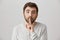 Close-up portrait of confident tired male with beard making shush gesture while standing over gray background. Man asks