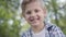 Close-up portrait of cute handsome boy in checkered shirt looking into camera sitting in the park. Summertime leisure