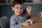 Close up of portrait of cute little boy eats delicious yogurt or cottage cheese from white container with spoon at table in dining