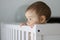 Close-up portrait of cute thoughtful baby staying in his baby cot holding the side and daydreaming