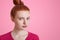 Close up portrait of freckled female with ginger hair bun, wears pink casaul sweater, looks confidently into camera, thinks about