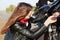 Close up portrait of girl motorist with long dark hair in jacket, trying to repair motorbike during trip, fixes some parts, has
