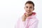 Close-up portrait of handsome creative male, blond hair and pink hoodie, hold hand near chin and smiling pleased, have