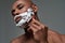 Close up portrait of handsome young african american man with foam applied on his face using steel razor for shaving