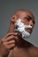 Close up portrait of handsome young african american man with foam applied on his face using steel razor for shaving