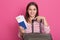 Close up portrait of happy European girl with suitcase, airplane tickets and passport isolated on rosy background, lady feels