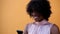 Close up and portrait of one young and beautiful afro American woman using her phone smiling and having fun with yellow wall at th