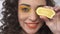 Close up portrait of playful young curly girl smiling and covering eye with round piece of ripe lemon. Slow motion