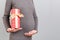 Close up portrait of pregnant woman in gray dress holding a gift box at gray background. Pregnancy celebration. Copy space