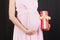 Close up portrait of pregnant woman in pink dress holding a gift box at black background. Pregnancy celebration. Copy space