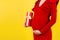Close up portrait of pregnant woman in red dress holding a gift box at yellow background. Pregnancy celebration. Copy space