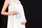 Close up portrait of pregnant woman in white dress holding blue socks for a baby boy at black background. Future child expectation