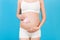 Close up portrait of pregnant woman in white underwear holding pink socks for a baby girl at blue background. Waiting for a child
