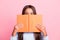 Close-up portrait of pretty funny brown-haired girl reading hiding behind book isolated over pink pastel color