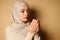 Close-up portrait of a serene young Arab woman in hijab praying. Religious beautiful woman in strict attire praying on beige