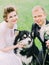 The close-up portrait of the smiling newlyweds petting the dog in the green field.