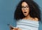 Close up portrait of stupefied young woman with Afro hairstyle, keeps mouth widely opened, holds smartphone, receives shocking