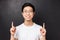 Close-up portrait of successful pleased young asian male entrepreneur, pointing fingers up and looking camera with