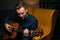 Close-up portrait of talented guitarist singer male playing acoustic guitar sitting on armchair in dark living room.