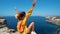 Close up portrait of taned traveler woman sitting on top rock beach with raised hands, bright yellow dress and hair