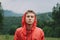 Close up portrait of a thoughtful young tourist man wearing a red raincoat and hood seriously looking at the camera, feeling calm