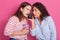 Close up portrait of two surprised cute young women using mobile phone together over pink background, attractive ladies hearing