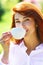 Close up portrait of a woman holding a hot coffee cup outdoors on the spring background. Red head girl drinking coffe.