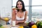 Close-up portrait of woman standing in kitchen, leaning on wooden table, having snack. Smiling girl maintains healthy