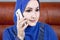 The close up portrait of a young attractive muslim woman in a blue dress sits on the sofa at home, talks on the white mobile phone