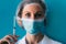 Close-up portrait of young female doctor in medical cap and mask, white gown and syringe on her hand on blue background.