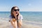 Close-up portrait of young girl with long hair standing near blue sea. She wears black sunglasses. She is surprised at