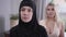 Close-up portrait of young Muslim woman in hijab looking at camera. Blurred modern Caucasian woman standing at the