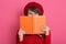 Close up portrait of young woman hides her face behind her notebook orange notebook, charming female wears red sweater, beret and