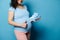 Close-up pregnant woman with big belly, shows thumb up, smiles at camera, holding newborn baby clothes, isolated on blue