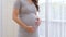 Close up Pregnant Woman standing in front of windows and stroking her big belly with love at cozy home,Pregnancy of young woman