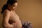 Close-up pregnant woman stroking her tummy, holding bouquet of purple blooming lilacs, isolated beige studio background