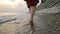 Close-up rear view of legs Caucasian girl walks along the water on the stone coast of the sea towards the setting sun at