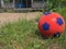 Close-up of a red soccer ball for children on the green grass in front of a country house. Porch of the house in the background.