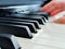 A close-up of a row of piano keys. In the blurred background, a pianist\'s hands can be seen practicing on the piano.