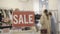 Close-up of Sale announcement in clothing store with blurred female shoppers choosing clothes at the background. Wealthy