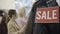 Close-up of Sale banner on pricey clothes hanging on rack with blurred beautiful Caucasian women choosing garment at the