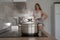 Close-up of a saucepan with boiling water on the stove. A young woman stands aside.