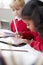 Close up of a schoolgirl sitting at desk next to a boy in an infant school classroom, using a tablet computer and stylus, selectiv