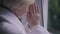 Close-up senior wrinkled hands together of grey-haired woman praying looking out the window indoors. Thoughtful