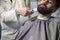 Close up of a serious brown haired businessman having his beard combed and trimmed in a barber shop.