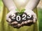 Close-up the shining 2023 year numbers calendar on organic soil in the hands while holding young green sprout plant.