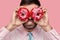 Close up shot of bearded businessman in light pink formal shirt covers eyes with two delicious doughnuts, clenches