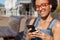 Close up shot of black smiling woman meloman enjoys radio online, holds mobile phone, connected to digital earphones